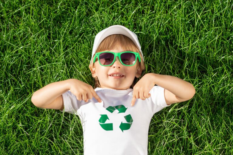 little boy lying in grass wearing green sunglasses, a white cap, and a white tshirt with the reduce reuse recycle sign on it
