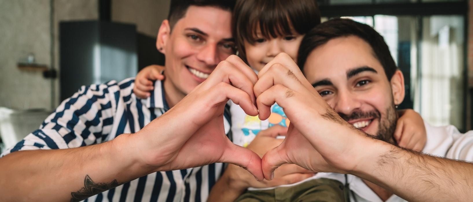 close up of two fathers with small child sitting between their heads on their shoulders, both are holding one hand up towards the camera and cupping them together to form a heart in front of child